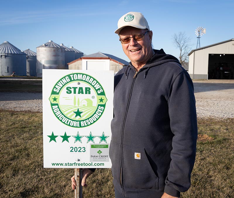 Farmer Steve Stierwalt with STAR field sign, with silver machine shed and bins in back.